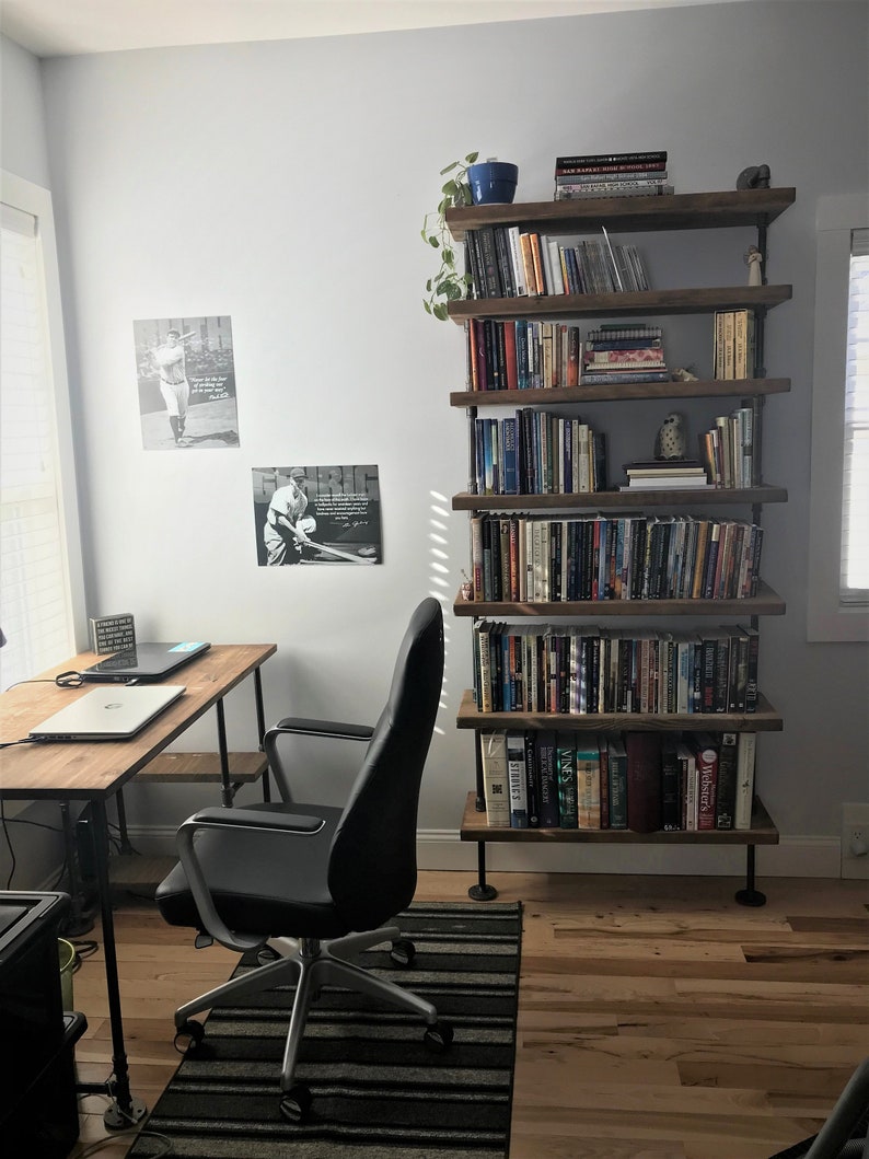 May include: A black office chair with wheels sits in front of a wooden desk with a laptop and a plant on a shelf. A tall, industrial-style bookshelf with multiple shelves holds books and decorative items. The bookshelf is made of wood and metal.