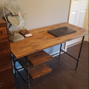 May include: A wooden desk with a black metal frame and three shelves. The desk has a laptop computer on the top shelf and a fan on the floor next to the desk.
