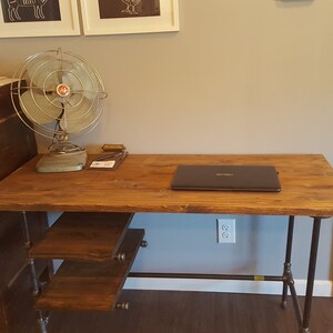May include: A wooden desk with a black laptop on the surface. The desk has a metal pipe frame and three wooden shelves. A vintage metal fan is on the surface of the desk next to the laptop. The desk is in a room with a file cabinet and a white door.