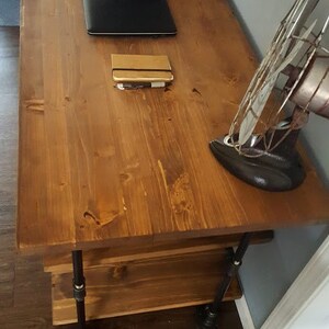 K&ouml;nnte beinhalten: Ein Holzschreibtisch mit dunkelbrauner Oberfl&auml;che und schwarzen Metallrohrbeinen. Ein Laptop, ein Notizbuch und ein Vintage-Ventilator stehen auf dem Schreibtisch.