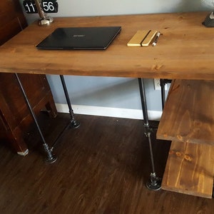 K&ouml;nnte beinhalten: Ein Holzschreibtisch mit schwarzen Metallrohrbeinen und zwei Regalen. Der Schreibtisch hat einen Laptop, einen Stift und ein Notizbuch auf der Oberseite.