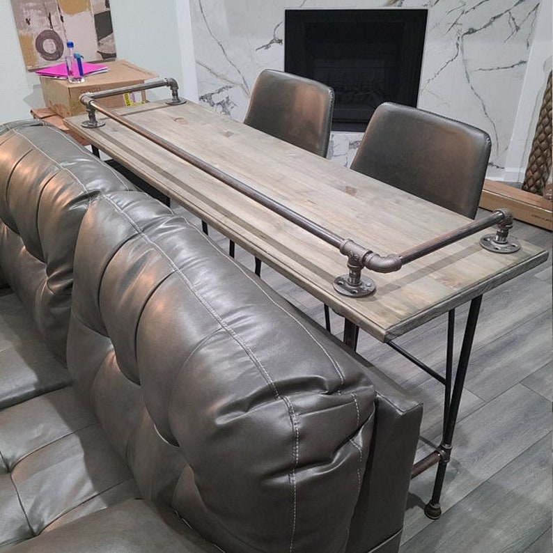 May include: A rustic wooden bar table with a metal pipe rail and black metal legs. The table is placed in front of a gray leather couch.