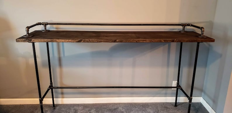 May include: A rustic wooden bar table with black metal pipe legs and a top rail. The table has a dark brown stain and is made of reclaimed wood.