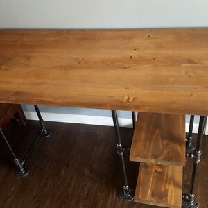 K&ouml;nnte beinhalten: Ein Holzschreibtisch mit einer braun gebeizten Platte und schwarzen Metallrohrbeinen. Der Schreibtisch hat zwei Regale unter der Platte.