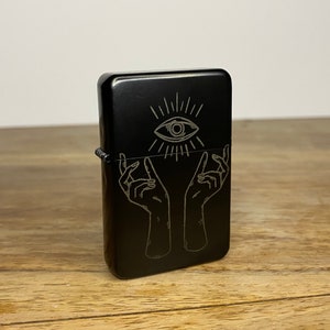 May include: Black lighter featuring a silver design of an eye and hands. The eye is at the top, with rays extending from it. Below the eye are two hands reaching upwards. The lighter is rectangular with a metal hinge.