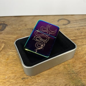 Pu&ograve; includere: Un accendino arcobaleno con una custodia argentata e un designo di un serpente avvolto attorno a una luna crescente. L'accendino &egrave; in una scatola argentata con un interno nero.