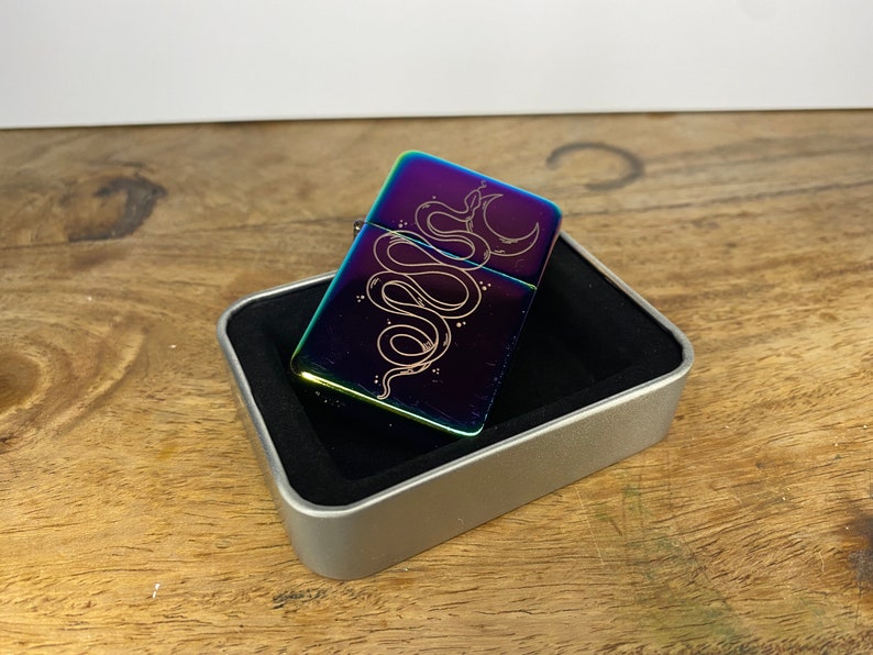 Pu&ograve; includere: Un accendino arcobaleno con una custodia argentata e un designo di un serpente avvolto attorno a una luna crescente. L'accendino &egrave; in una scatola argentata con un interno nero.