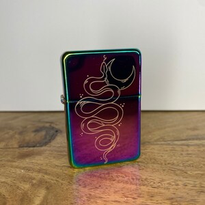 Pu&ograve; includere: Un accendino color arcobaleno con una cerniera argentata. L'accendino presenta un designo di un serpente avvolto attorno a una luna crescente.