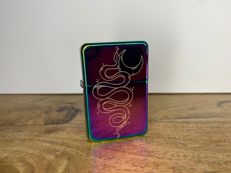 Pu&ograve; includere: Un accendino color arcobaleno con una cerniera argentata. L'accendino presenta un designo di un serpente avvolto attorno a una luna crescente.