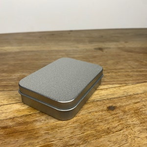Pu&ograve; includere: Una scatola di metallo argentata con una superficie testurizzata. La scatola &egrave; chiusa e si trova su una superficie di legno.