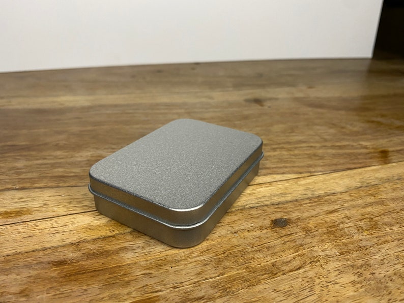 Pu&ograve; includere: Una scatola di metallo argentata con una superficie testurizzata. La scatola &egrave; chiusa e si trova su una superficie di legno.