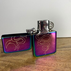 Pu&ograve; includere: Un accendino iridiscente arcobaleno con un designo a serpente inciso sul davanti e sul retro. L'accendino &egrave; aperto e la ruota della pietra focaia &egrave; visibile.