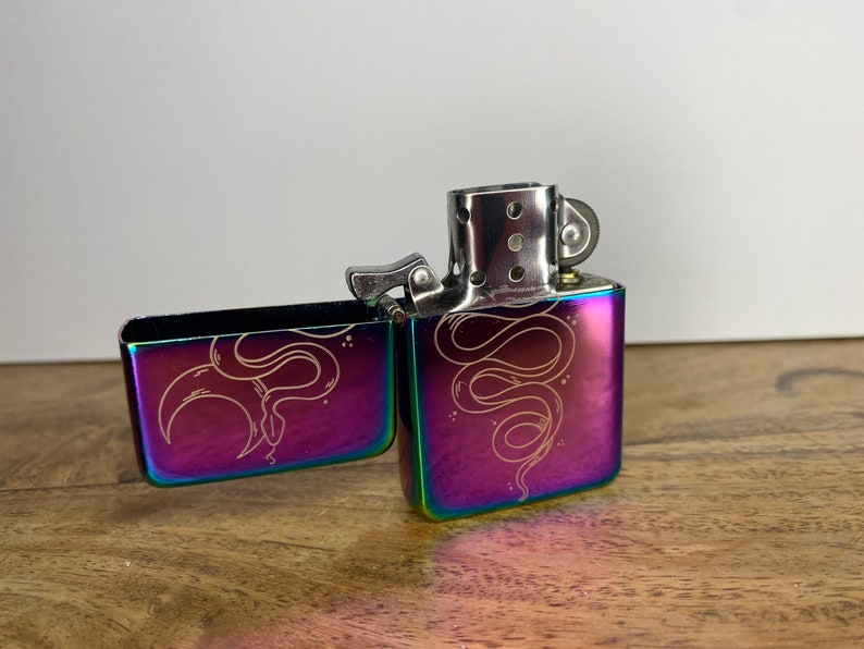 Pu&ograve; includere: Un accendino iridiscente arcobaleno con un designo a serpente inciso sul davanti e sul retro. L'accendino &egrave; aperto e la ruota della pietra focaia &egrave; visibile.