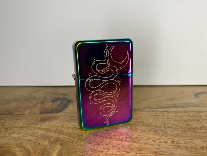 Pu&ograve; includere: Un accendino iridiscente arcobaleno con un designo inciso a laser di un serpente avvolto attorno a una luna crescente.