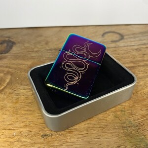 Pu&ograve; includere: Un accendino arcobaleno iridescente con una custodia argentata e un designo di un serpente e una luna crescente inciso sul davanti.