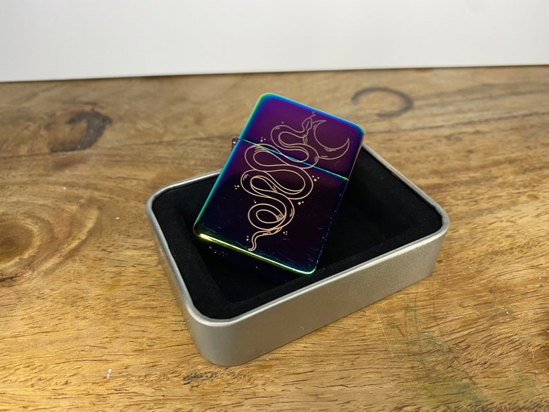 Pu&ograve; includere: Un accendino arcobaleno iridescente con una custodia argentata e un designo di un serpente e una luna crescente inciso sul davanti.