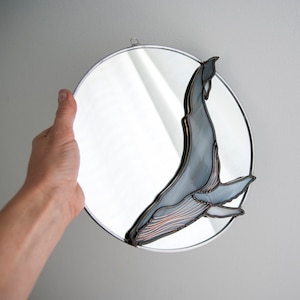 May include: A round mirror with a silver frame features a stained glass whale design. The whale is blue and gray with copper accents. The mirror is being held by a hand.