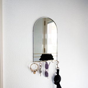 May include: A silver arched mirror with a shelf and hooks for hanging keys, sunglasses, and a leash. The mirror is mounted on a white wall.