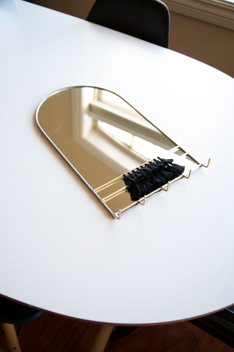 May include: A silver arched mirror with a black tassel hanging from the bottom. The mirror has three hooks on the bottom.