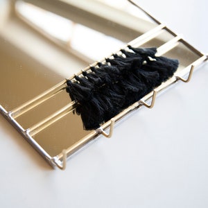 May include: A gold-toned metal mirror with three hooks and a black tassel trim. The mirror is rectangular and has a reflective surface.