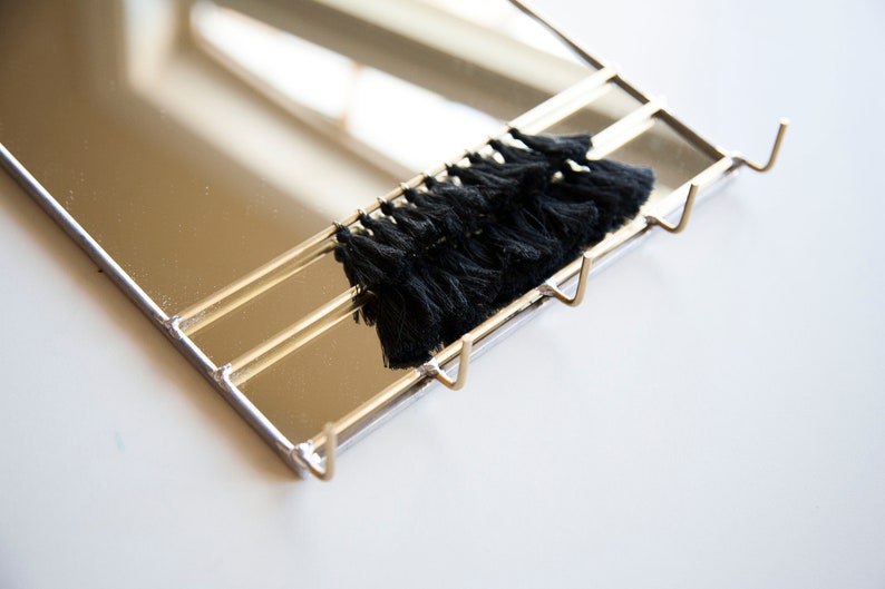 May include: A gold-toned metal mirror with three hooks and a black tassel trim. The mirror is rectangular and has a reflective surface.