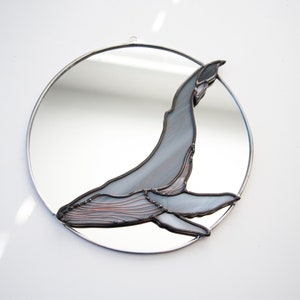 May include: A round mirror with a stained glass whale design. The whale is gray and has a copper wire outline. The mirror is framed in silver.