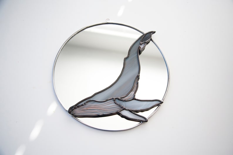 May include: A round mirror with a stained glass whale design. The whale is gray and has a copper wire outline. The mirror is framed in silver.