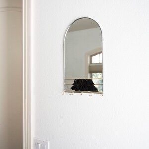 May include: A silver arched mirror with a black fringe shelf and gold hooks underneath. The mirror is mounted on a white wall.