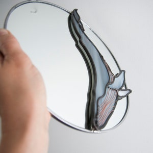 May include: A round mirror with a stained glass whale design. The whale is made of blue, gray, and copper glass. The mirror is hanging on a white wall.