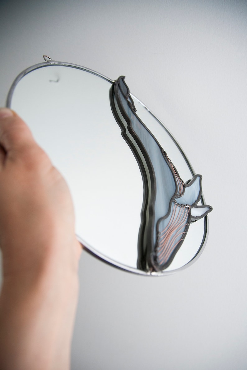 May include: A round mirror with a stained glass whale design. The whale is made of blue, gray, and copper glass. The mirror is hanging on a white wall.