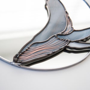 May include: A round mirror with a stained glass whale design. The whale is made of blue and gray glass with copper wire accents.