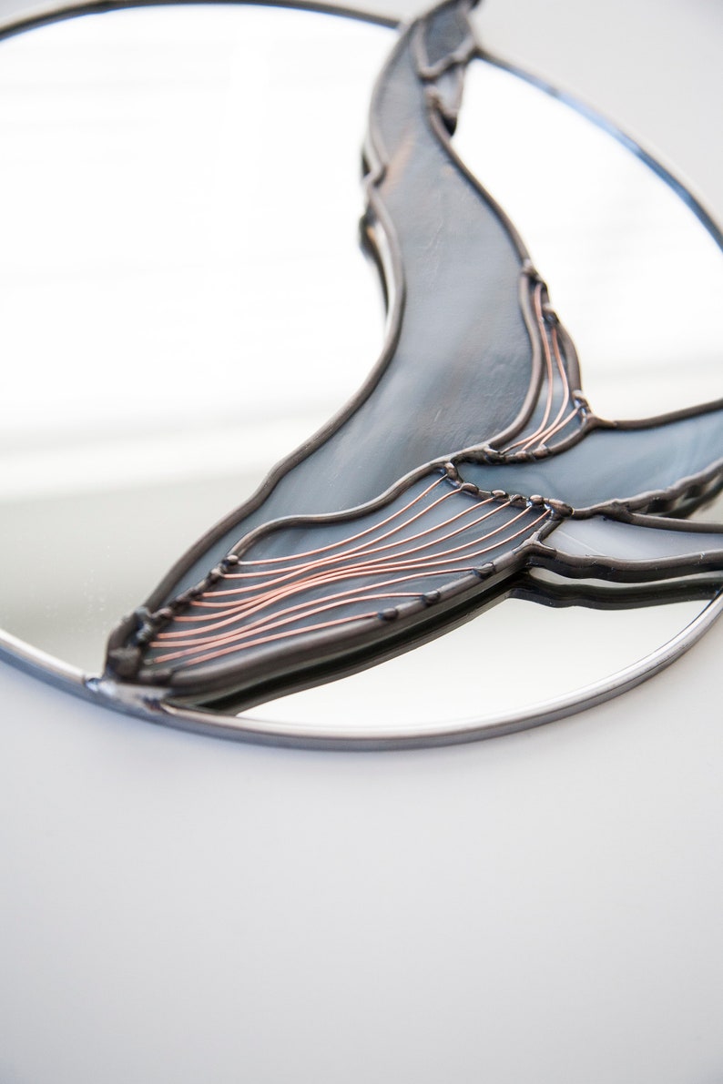May include: A round mirror with a stained glass whale design. The whale is made of blue and gray glass with copper wire accents.