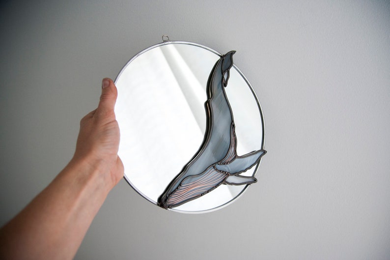 May include: A round mirror with a silver frame features a stained glass whale design. The whale is blue and gray with copper accents. The mirror is being held by a hand.