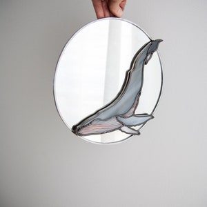 May include: A round mirror with a stained glass whale design. The whale is blue and gray with a copper wire outline. The mirror is hanging from a white wall.