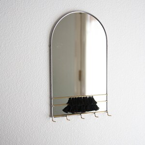 May include: A silver arched mirror with a gold shelf and hooks. The mirror has a black tassel hanging from the shelf.
