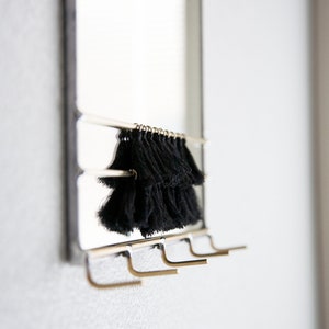 May include: A silver mirror with a black tassel hanging from the top. The mirror has four gold hooks on the bottom.