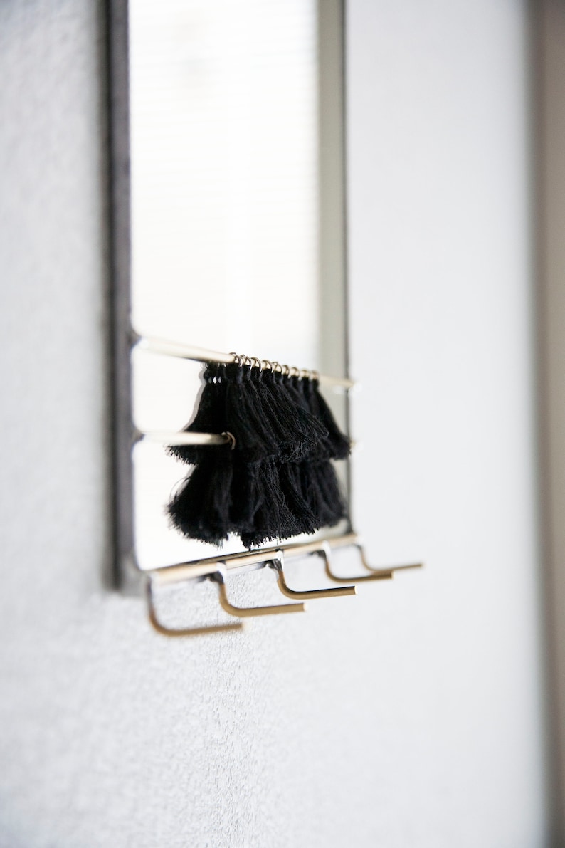 May include: A silver mirror with a black tassel hanging from the top. The mirror has four gold hooks on the bottom.