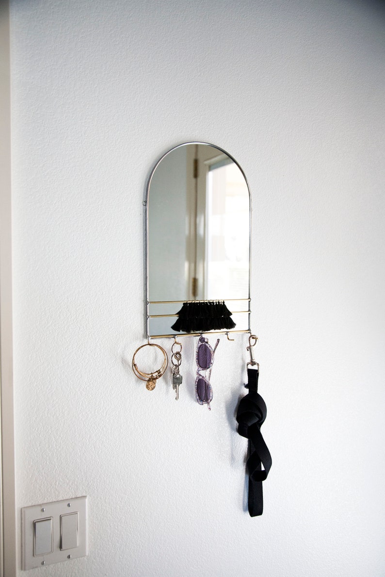 May include: A silver arched mirror with a shelf and hooks for hanging keys, sunglasses, and a leash. The mirror is mounted on a white wall.
