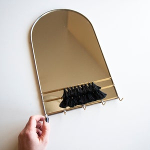 May include: A gold-framed arched mirror with a black tassel trim and three hooks underneath. The mirror is hanging on a white wall.