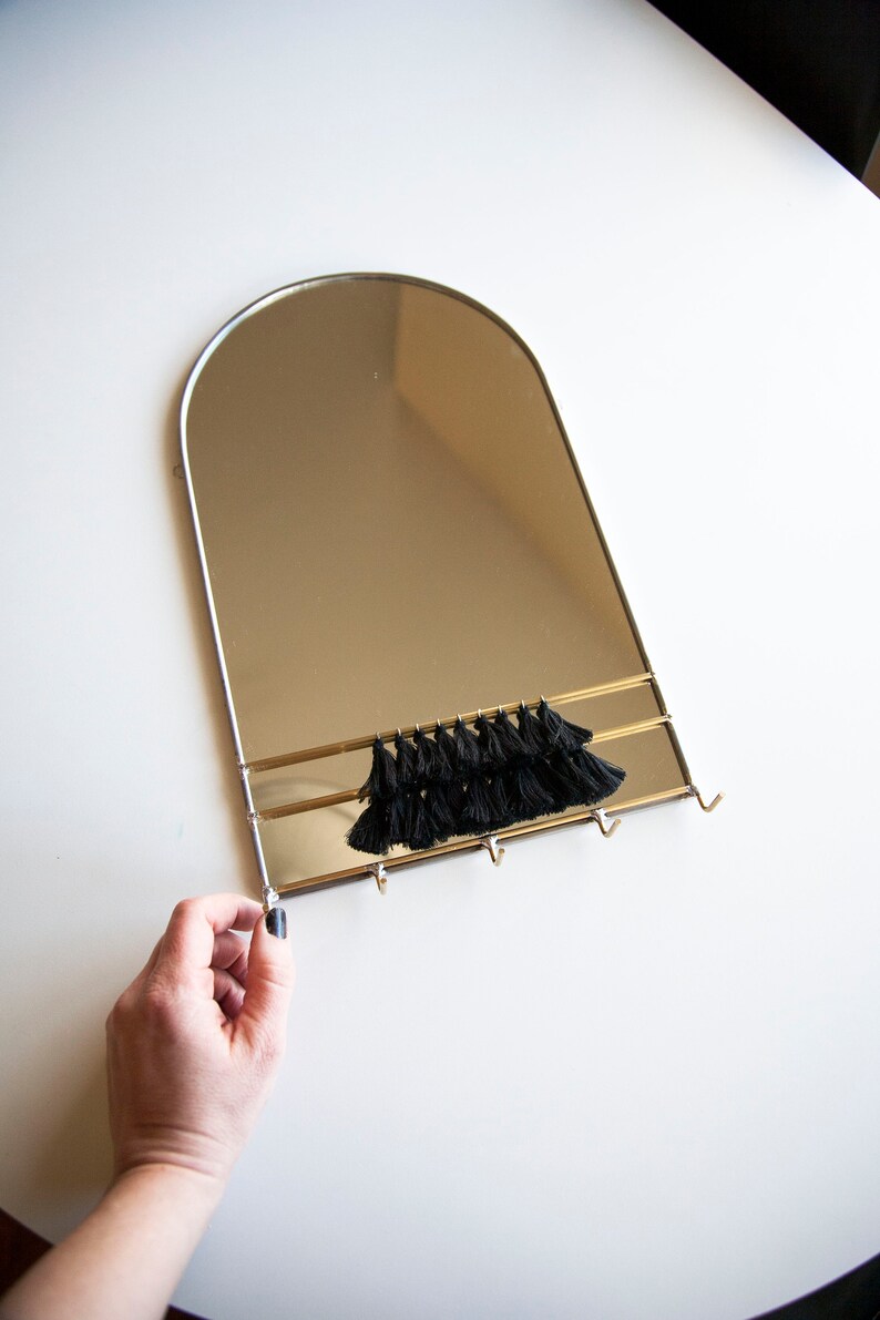 May include: A gold-framed arched mirror with a black tassel trim and three hooks underneath. The mirror is hanging on a white wall.