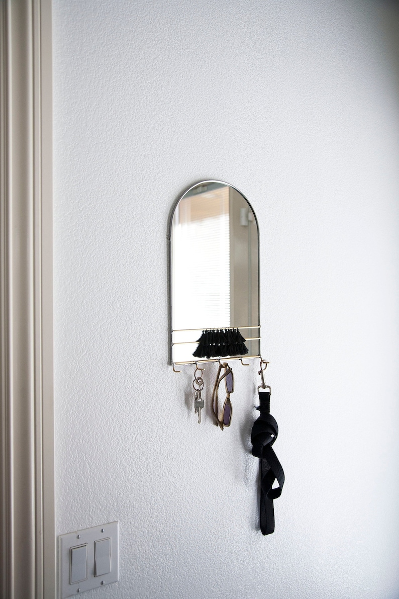 May include: A silver arched mirror with a shelf and hooks for hanging keys, sunglasses, and a black strap. The mirror is mounted on a white wall.