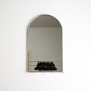 May include: A silver arched mirror with a brass shelf and black tassels hanging from the shelf. The mirror is mounted on a white wall.