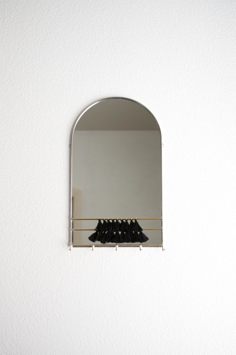 May include: A silver arched mirror with a brass shelf and black tassels hanging from the shelf. The mirror is mounted on a white wall.