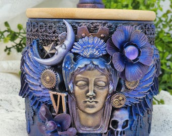 May include: A decorative jar with a wooden lid and a dark blue base. The jar is adorned with sculpted details including a face, wings, flowers, a moon, and gears. The lid has a dark purple knob.