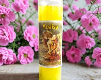 May include: A tall cylindrical glass candle filled with bright yellow wax. The label features the name Anaisa in bold yellow letters above an illustration of a person with long hair wearing a flowing orange garment. The text reads Mystical Candle.