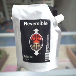 May include: A white flexible pouch containing Mystical Bath liquid. The black label features the word Reversible, an illustration of a face with an inverted reflection, and instructions to shake before use in a gallon of water for nine minutes.