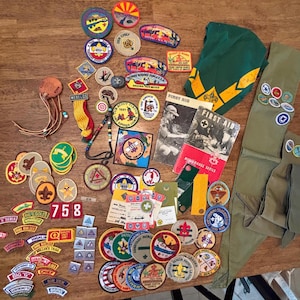 May include: A collection of vintage Boy Scout patches, badges, and ephemera. The assortment includes circular and rectangular patches with various designs, colors, and text, along with a green neckerchief and a First Aid booklet.
