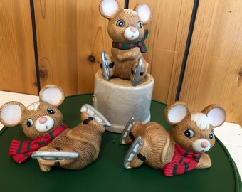 May include: A collection of ceramic mouse figurines, each with brown fur, white ears, and blue eyes. The mice wear red and green scarves and silver ice skates. One mouse sits atop a small, beige cylinder.