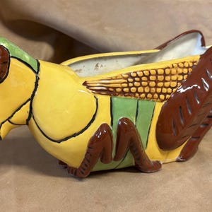 May include: A ceramic grasshopper-shaped planter. The planter is painted with bright colors, including yellow, green, and brown. The grasshopper has large eyes and detailed wings. The planter is designed to hold small plants or flowers.