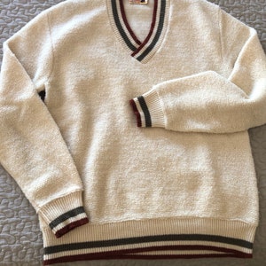 May include: Cream-colored V-neck sweater with a textured knit. The collar, cuffs, and hem feature stripes in dark green, burgundy, and cream. The sweater is hanging on a black hanger.